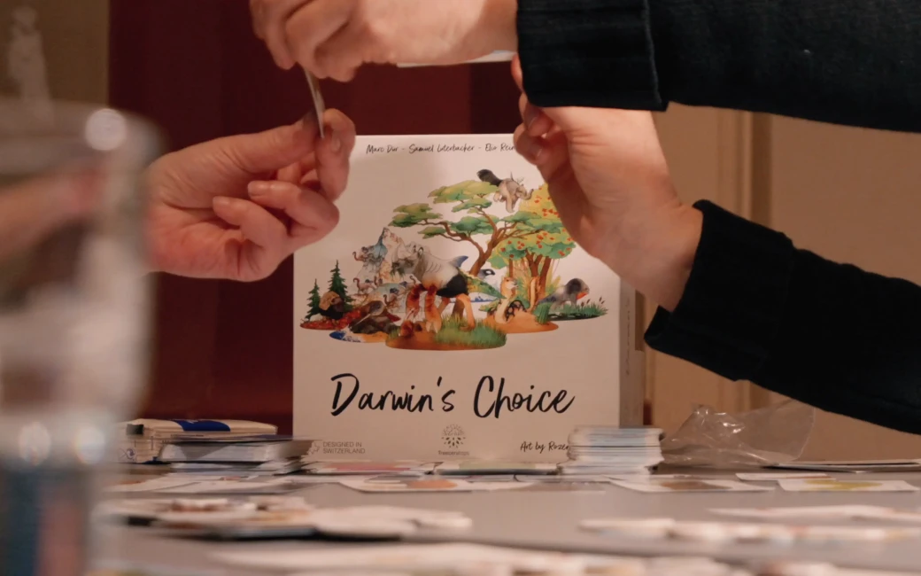 Im Hintergrund steht die Schachtel des Spieles “Darwin´s Choice”. Davor sind die Hände einiger Teilnehmender zu sehen.