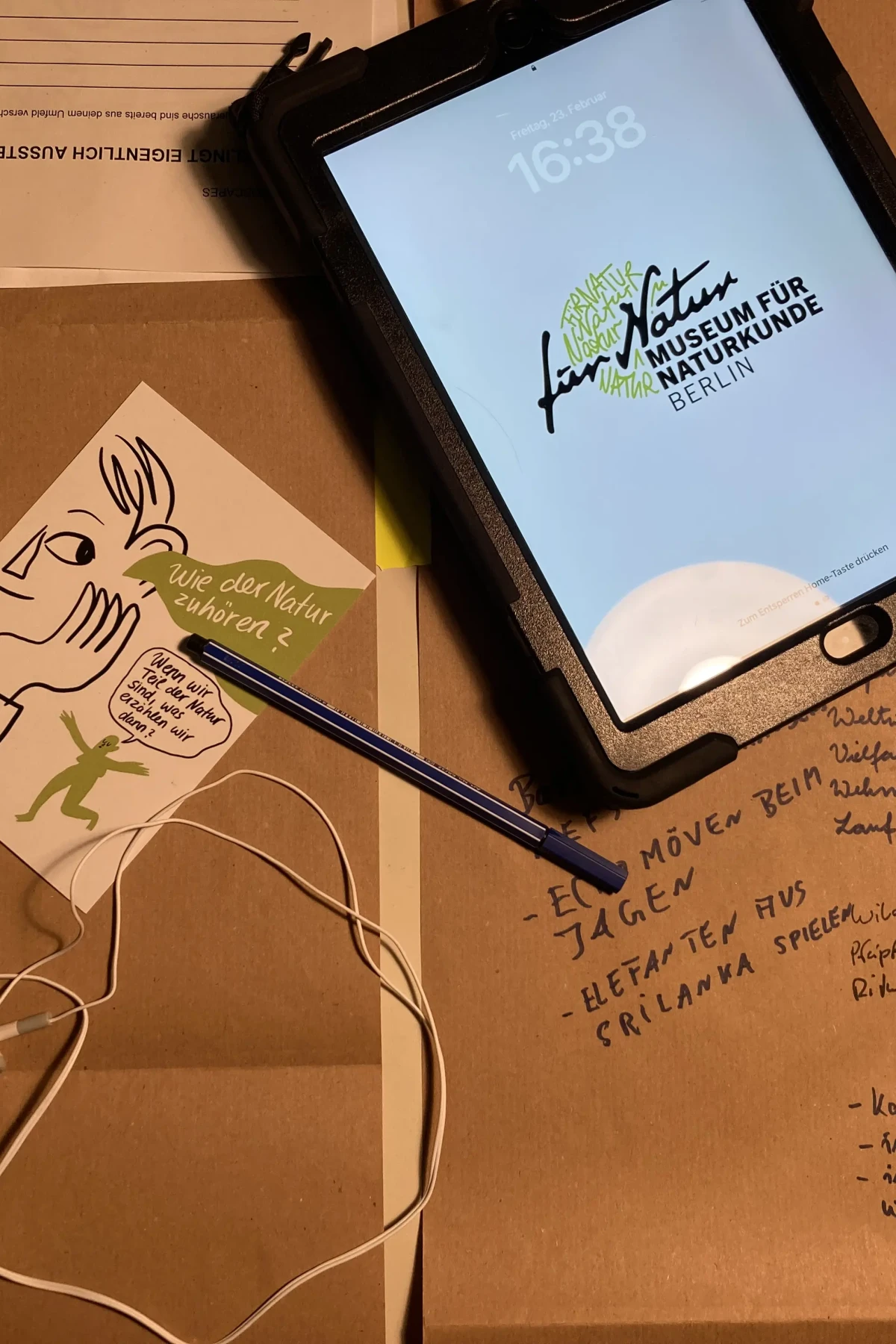 Abbildung des Arbeitsprozesses. Zu sehen sind Mitschriften, Postkarten, Papiere, Stifte, Kopfhörer sowie ein Tablet.